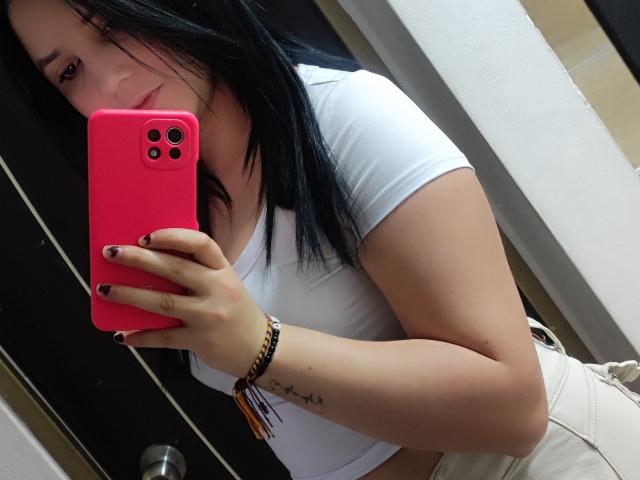 Kkatalina with black hair, white tee, and beige pants, holding pink phone, showing tattoo on left wrist, and smiling at mirror selfie. Boobs visible under tight shirt.