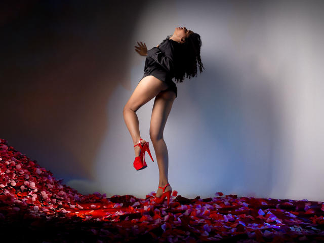 ZoeJonees con una camisa negra, sin ropa en la parte inferior, tacones rojos altos, de pie sobre pétalos de rosa rojos, inclinada hacia atrás con los ojos cerrados, cabello negro largo, piel oscura, pose sexy..