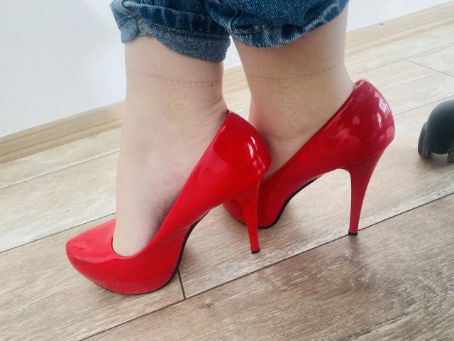 Steisii's shiny red high heels with stiletto heels, standing on wooden floor, legs in blue jeans, cuffed at ankles, visible tan socks with faint patterns.