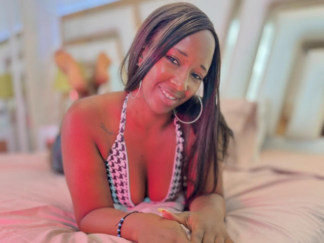 EmmaGrace69 with dark brown hair, wearing a black-and-white houndstooth bikini, has big tits and a tattoo on her left shoulder, smiling on a bed with pink sheets.