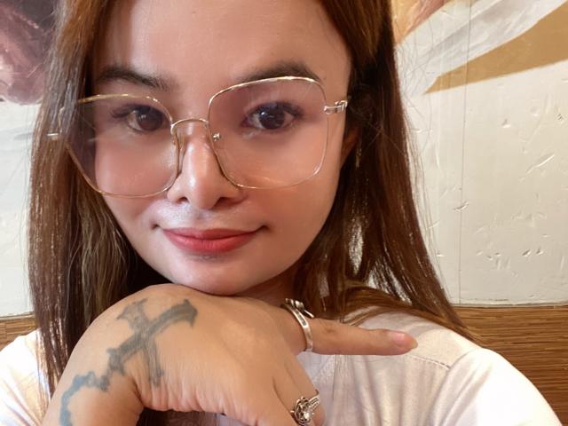 LexiBest69 has light brown skin, long brown hair, and wears large clear glasses. She's smiling with a tattoo on her right hand and a ring on her index finger.