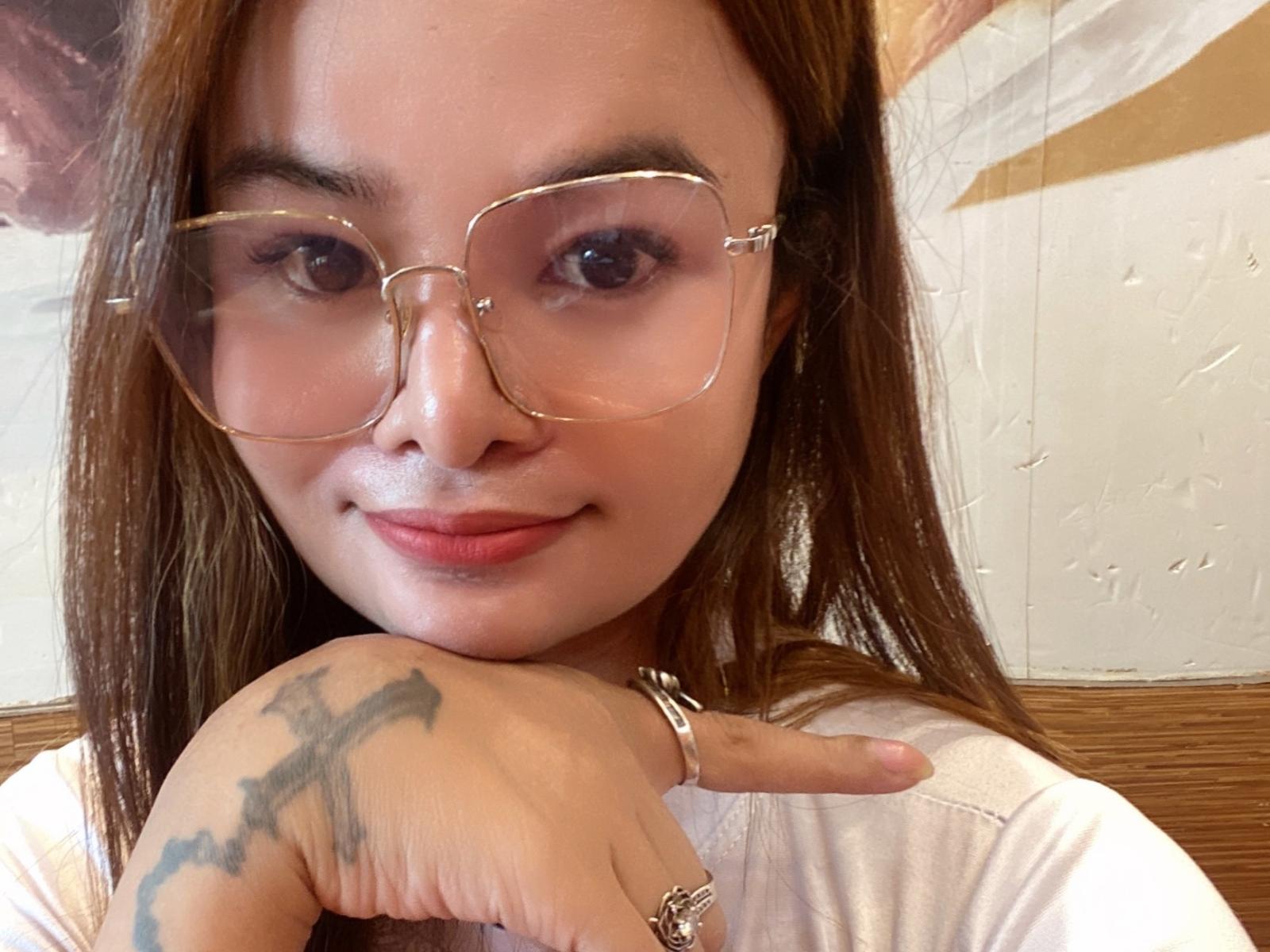 LexiBest69 has light brown skin, long brown hair, and wears large clear glasses. She's smiling with a tattoo on her right hand and a ring on her index finger.