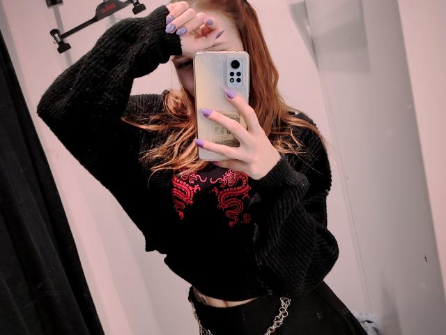 MandyBrooks with red hair, black sweater, red graphic tee, black skirt, holding phone covering face, purple nails, mirror selfie, wearing black cardigan, chain belt, in a bathroom.