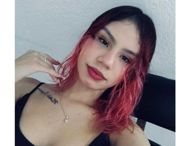SussyGray has red-dyed hair, dark eyebrows, and red lipstick. She's wearing a black top with visible tits, a tattoo on her right collarbone, and a necklace.