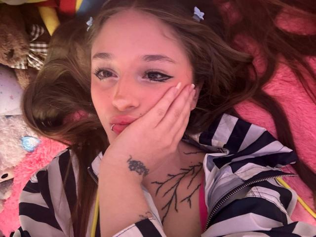 UwuMaddiee with long brown hair, blue eyes, and black eyeliner, lying on pink blanket, hand on face, striped jacket, pink bra, nose piercing, tree tattoo on chest.