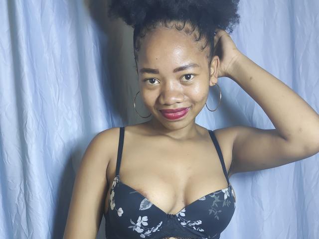 Aangela with dark skin, curly black hair, wearing a black floral bra, smiling, red lipstick, hoop earrings, hand in hair, standing against a white curtain.
