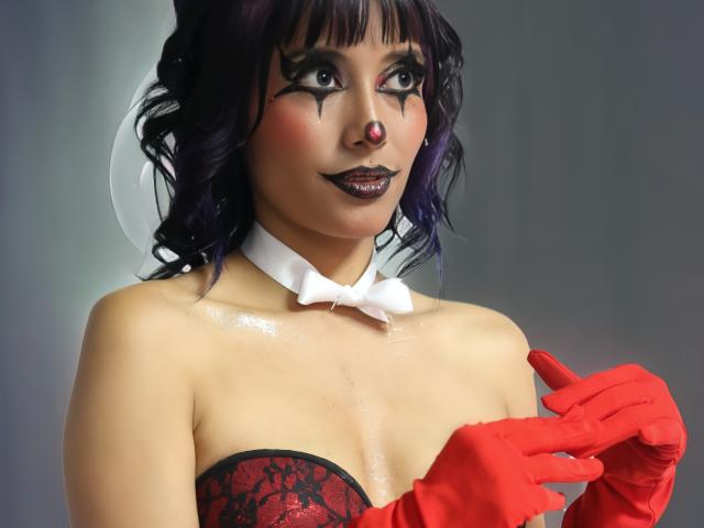AbyColeman with dark purple hair, black makeup, red nose, black lipstick, white bowtie, red gloves, and red lace top, showing off shiny tits, holding gloves.