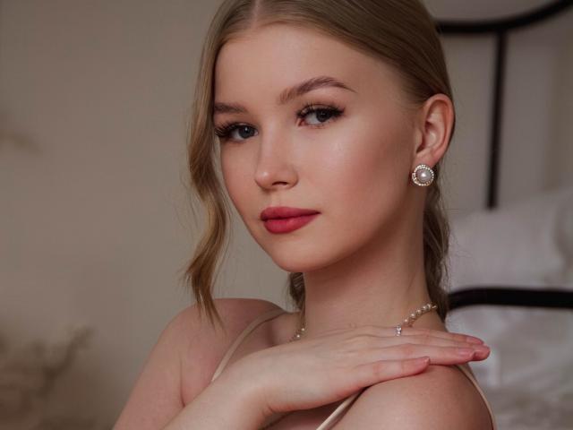 NicoleBrian has blonde hair, fair skin, blue eyes, red lipstick, and is wearing diamond earrings with a delicate necklace. Her hand is on her shoulder.