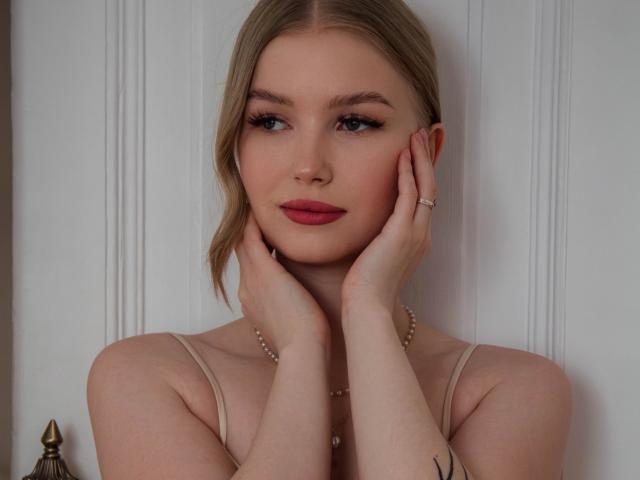 NicoleBrian has pale skin, blonde hair, and red lipstick. She's touching her face with hands, wearing a pearl necklace and a beige top. Her tits are small, and she's cute.