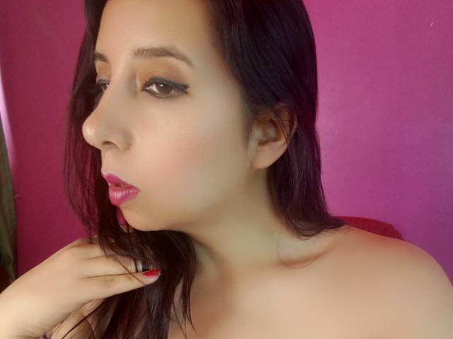 SublimeMature has fair skin, dark straight hair, pink lipstick, and red nails, with a subtle, sexy, pouty expression, and a hand touching her neck.