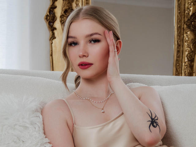 NicoleBrian with blonde hair, pink lipstick, and a spider tattoo on her right arm, wearing a beige dress, touching her face, pearl necklace, sitting on a white couch.