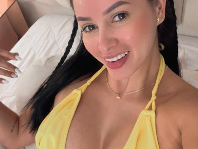 EmilyParkerr, con un top da bikini giallo, sorride alla fotocamera, mostrando il seno, indossa una collana d'oro e ha i capelli intrecciati, sdraiata su un letto..