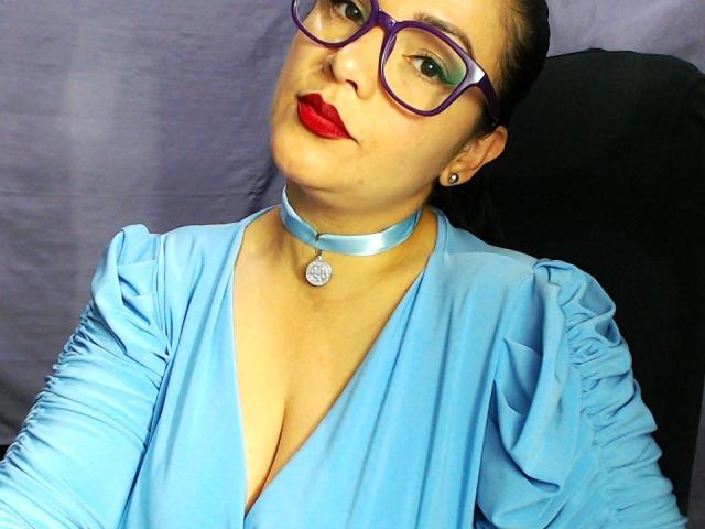 NatalyBlaine with purple glasses, red lipstick, blue dress, and cleavage, wearing a silver choker with a ring, sitting on a black surface, looking up.