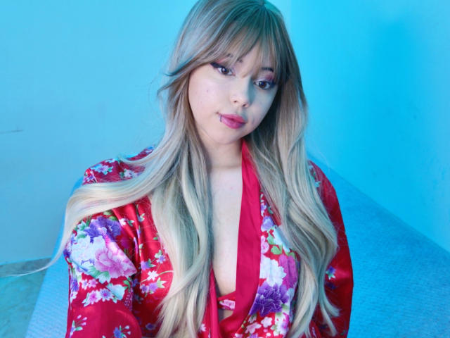 Arieyaara with long blonde hair wearing a red floral kimono, showing a cleavage, sitting in blue light, looking at the camera with a slight smile.