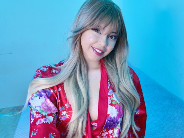 Arieyaara with long blonde hair, red floral kimono, and a smile, showing her tits in a blue-lit setting. She's cute and sexy, with a playful expression.