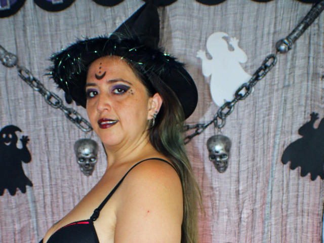 MaraLeon with green eyes, black witch hat, dark makeup, and green hair, wearing a black bra, showing her tits, and skull earrings, smiling at the camera.