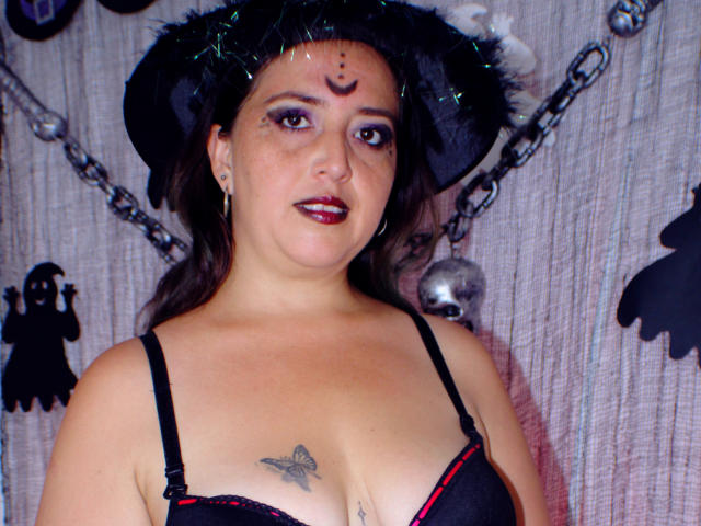 MaraLeon with dark hair, black hat, red lipstick, black bra, tattoo on chest, nose ring, and chain, standing, showing tits, and a cute face, in a Halloween vibe.
