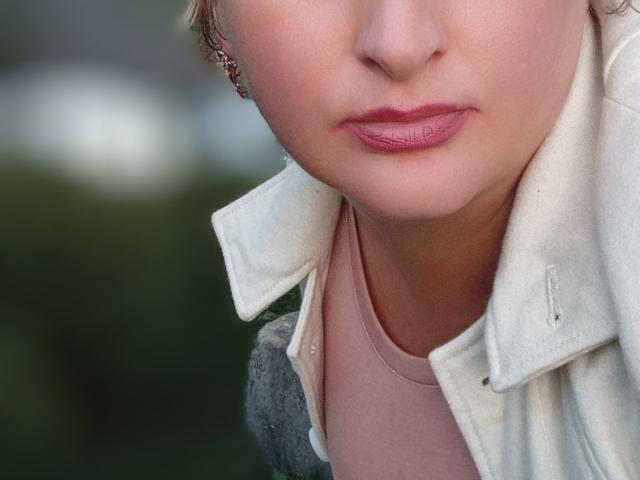 Het gezicht van KelliRedhaer, met roze lippenstift en licht open roze lippen, draagt een witte jas over een roze shirt, waarbij haar neus en bovenlip zichtbaar zijn..