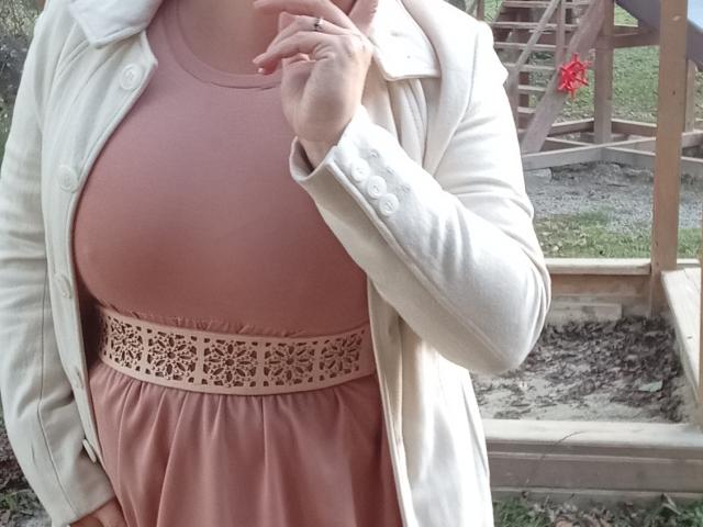 KelliRedhaer's pink dress with lace belt, white coat, and hand touching neck. Boobs are visible, slightly covered by coat. Her fingers are adorned with a simple ring.