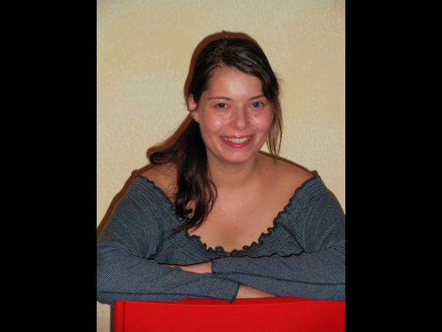 Chiara177-hot with dark brown hair, smiling, wearing a gray off-shoulder top, arms crossed, red cloth in front, showing her bare tits, against a yellow wall.