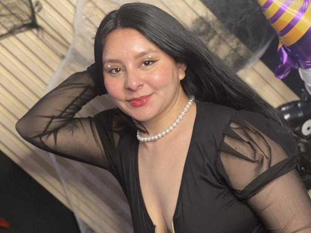 EmaJames mit langem, schwarzem Haar, trägt ein durchsichtiges schwarzes Kleid, eine Perlenkette und roten Lippenstift, lächelt in die Kamera und zeigt ihre Brüste in einem tief ausgeschnittenen Kleid..