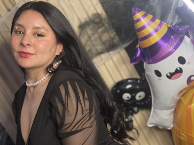 EmaJames with long black hair, wearing a black sheer top, pearl necklace, and red lipstick, smiling at the camera, next to Halloween decorations including a purple hat ghost.