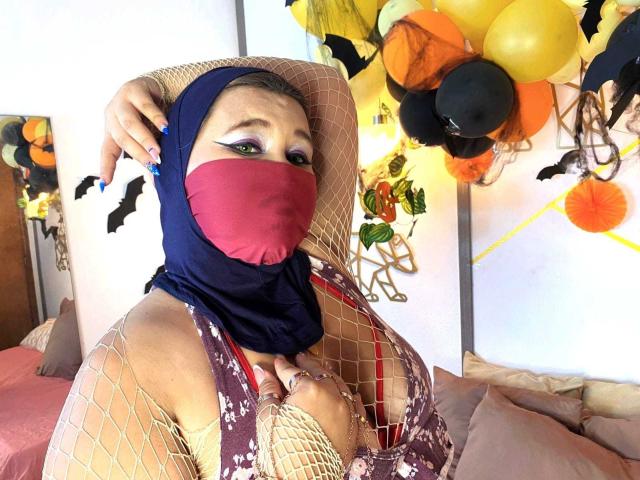 SalmaAzaman with big tits, wearing a red mask, blue hijab, fishnet top, and beige hat, surrounded by Halloween balloons, has green eyeshadow and blue nails.