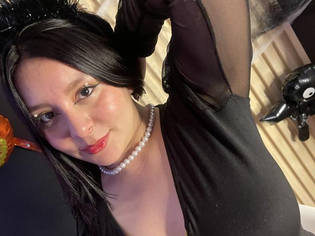 EmaJames, con cabello negro, vistiendo una blusa negra transparente, un collar de perlas y orejas de gato negras, sonriendo con lápiz labial rojo, con los brazos levantados, mostrando sus senos, y mirando a la cámara..