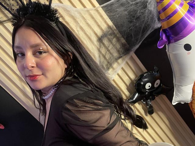EmaJames with black spiky crown, long black hair, sheer black top, pink lips, pearl necklace, looking over shoulder, black cat balloon, purple striped balloon, white balloon.