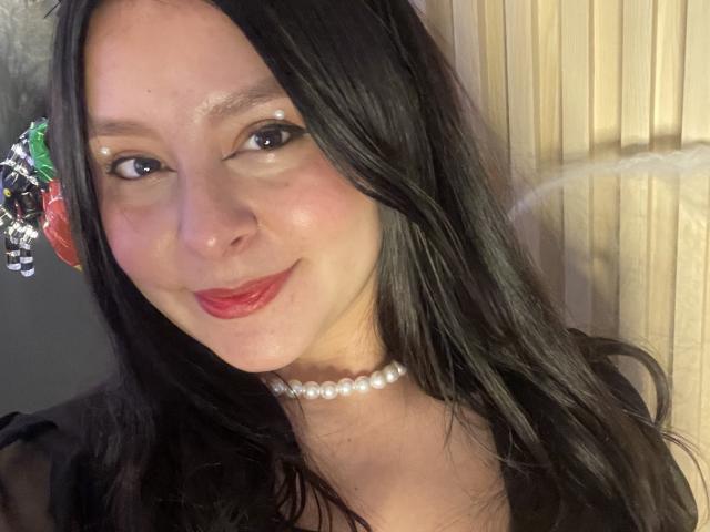 EmaJames has pale skin, black wavy hair, red lipstick, and a pearl choker. Her eyes are lined with glitter, and she's smiling softly. Boobs are not visible.