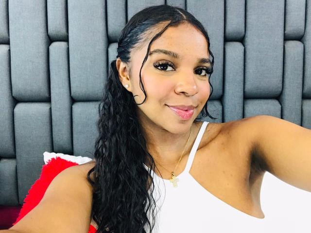 LisaMendez with black curly hair, brown skin, small tits, white tank top, gold necklace, puffy gray headboard, red pillow, and a slight smile. She's making a cute face.