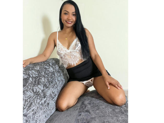 Emyi with long black hair, smiling, wearing a white lace lingerie top, black skirt, and white lace panties, kneeling on a gray textured couch. Her tits are visible.