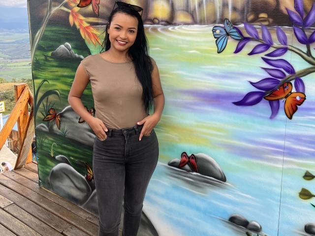 Emyi with long black hair, brown t-shirt, and gray jeans, standing with hands in pockets, smiling, against colorful graffiti wall, wooden deck, and outdoor setting.