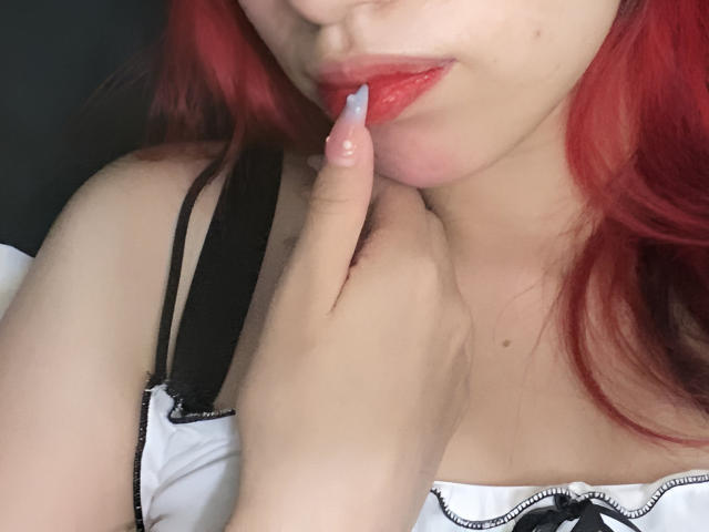 MilyRosie, com cabelo ruivo, mordendo o dedo com seus lábios vermelhos brilhantes, vestindo uma blusa branca com alças pretas, sua mão perto do pescoço, mostrando um pequeno corte no pulso..