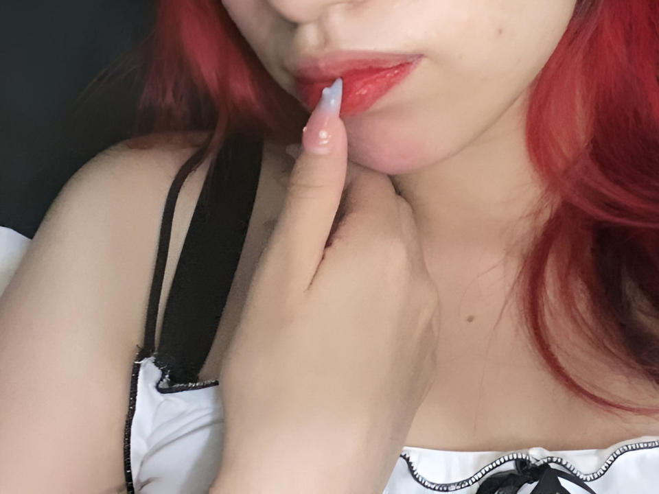 MilyRosie, con cabello rojo, mordiéndose el dedo con sus brillantes labios rojos, vistiendo una blusa blanca con tirantes negros, su mano cerca de su cuello, mostrando un pequeño corte en su muñeca..