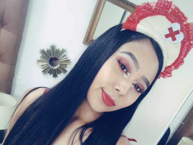 AliciaRichie with long black hair, red lipstick, and heavy makeup, wearing a red and white nurse hat, smiling at the camera. Boobs visible under a thin top.