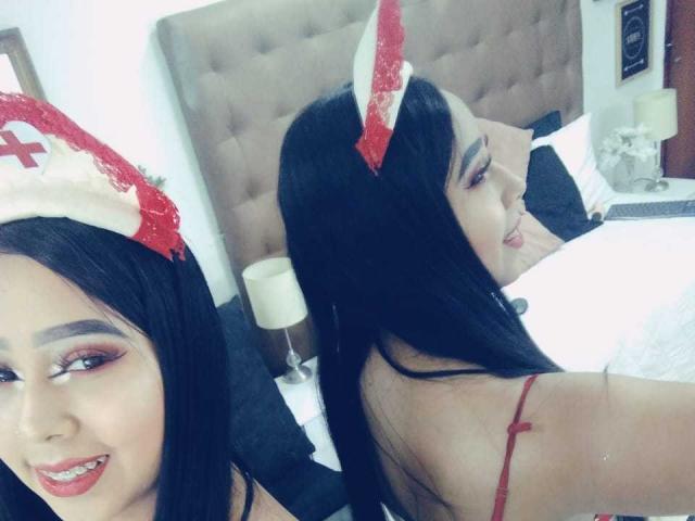 AliciaRichie with black hair, wearing a red cross nurse hat, smiling at the camera, showing red lipstick and eyeliner, reflected in a mirror, red strap visible.