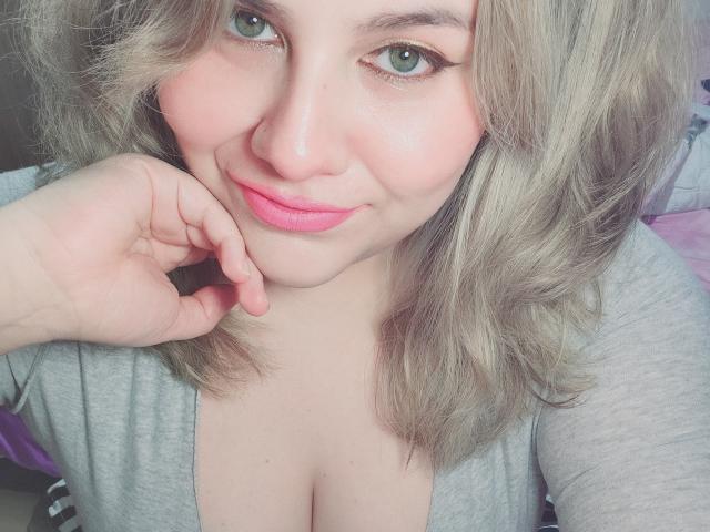 Akkanee, con i capelli grigi chiari, gli occhi verdi, il rossetto rosa e un leggero sorriso, appoggia il mento sulla mano, indossando una maglietta grigia che mette in mostra il seno..
