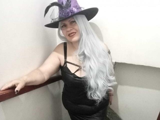 LesbiaThreeBebes con cabello largo y plateado-blanco, vestido de tirantes negros y un sombrero de bruja morado con plumas negras, sonriendo, apoyada en una barandilla roja, mostrando sus pechos y un rostro adorable..