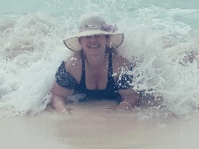 LesbiaThreeBebes com seios grandes, sorrindo, vestindo uma blusa preta, agachada nas ondas da praia, usando um chapéu de palha, com água espirrando ao redor dela..