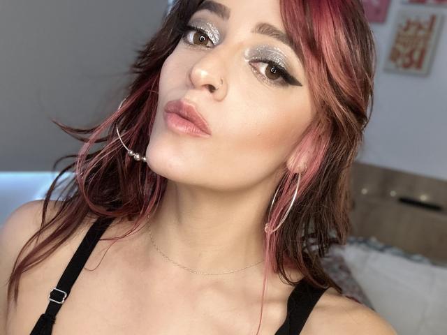 JaneRoa with red-pink hair, hoop earrings, nose ring, and silver eyeshadow, pouting with plump lips, wearing a black top, and a delicate chain necklace.