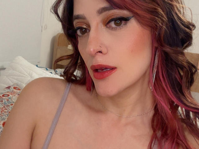 JaneRoa has pink-red hair, wearing red lipstick, winged eyeliner, and a nose ring, with a silver necklace and long earrings, showing a bit of her tits in a selfie.
