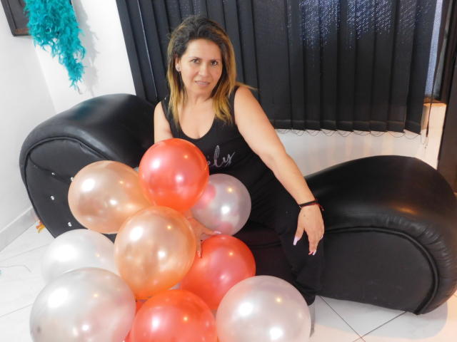 LesbiaThreeBebes, uma mulher de meia-idade com cabelo castanho, sentada em um sofá de couro preto, vestindo uma regata preta e calças pretas, segurando balões vermelhos e prateados, sorrindo..