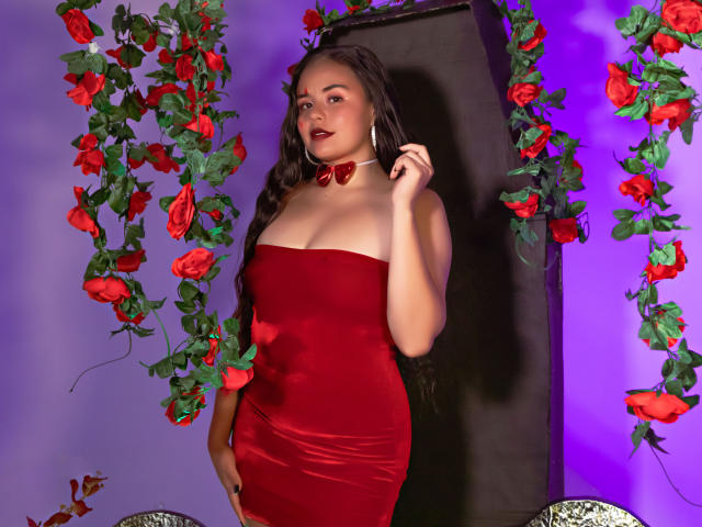 ArielleWilde with long brown hair, wearing a red velvet strapless dress, red bow choker, and large hoop earrings, standing in front of black wall with red rose garlands.
