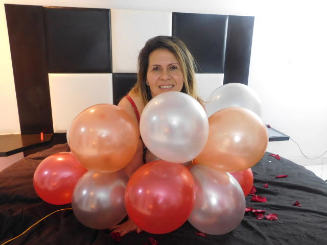 LesbiaThreeBebes sonriendo, sosteniendo un ramo de globos rosas, rojos y plateados, sentadas en una cama negra con pétalos de rosa..