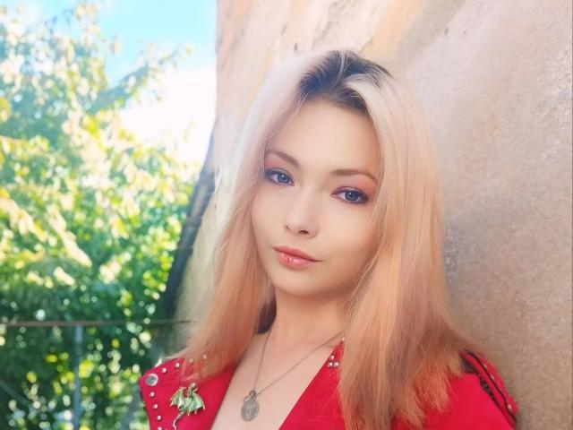 DevilMistress hat blondes Haar, blaue Augen und rosafarbenen Lippenstift. Sie trägt eine rote Jacke mit einer kleinen Blume und einer Halskette und blickt mit einem leichten Lächeln direkt in die Kamera..
