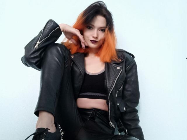DevilMistress with orange-to-black ombre hair, black leather jacket, black sports bra, black pants, and black boots, sitting with one knee up, hand on chin, looking at camera.
