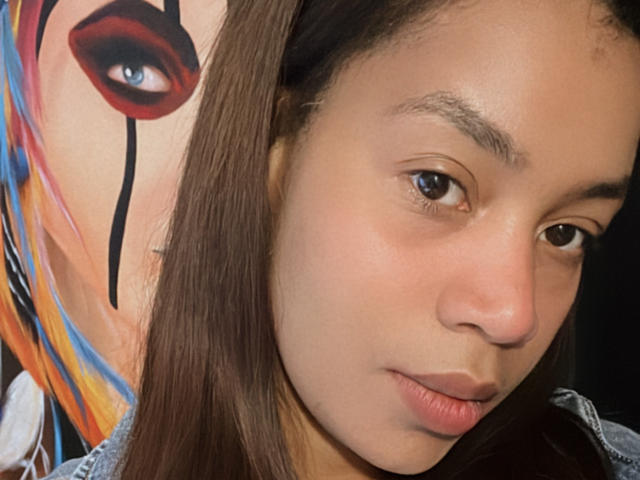 AlexaBigAss69's face with brown eyes, light brown skin, and long brown hair. A colorful painting of a woman with red lipstick and black eye makeup is in the background.