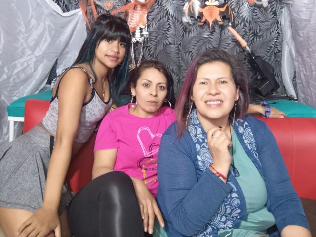 Tres Lesbias y tres bebés sentados juntos. Una con cabello negro y pantalones cortos grises, una con cabello castaño y una camisa rosa, una con cabello morado y un cárdigan azul. Todas sonriendo..
