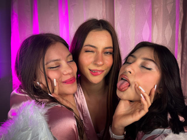 VictoriaAngels with three women, pink velvet, one winking, one tongue out, all with makeup, hands on faces, pink and purple lighting, one with nose ring, cozy and playful.